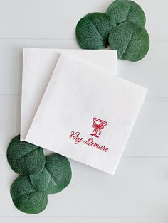 two white napkins with red lettering on them next to some green leaves and flowers