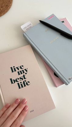 a woman's hand holding an open notebook next to a stack of books on a table