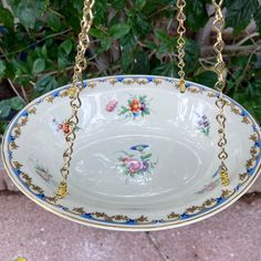 a bowl hanging from a chain with flowers and leaves on the bottom, in front of a bush