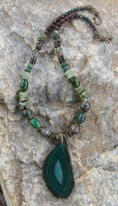 This attractive necklace is composed of many types of stones and beads:  there are 4 large tibetan line agate beads; 4 large green oval jasper gemstone beads; prehnite large chip/disc gemstone beads; fancy jasper beads; serpentine jasper beads; brown jade beads; Smokey Quartz beads; along with bronze pewter spacer beads and a hook claps, all of which are lead and nickel free. The pendant is 3 inches long and 1 1/2 inches wide, with crystals in the middle, framed by dark and light swirls of green and a few brown swirl lines.  The necklace is 20 1/4 inches long. Luxury Agate Beaded Necklace With Natural Stones, Luxury Beaded Agate Necklaces, Luxury Long Agate Necklace, Agate Necklace Green, Luxury Green Agate Beaded Necklaces, Green Agate Jewelry With Natural Stones, Spiritual Green Agate Jewelry, Earthy Agate Beaded Necklaces With Natural Stones, Green Agate Stone Jewelry