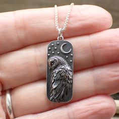 a hand holding a silver dog tag with an owl on it's back and the moon in the background