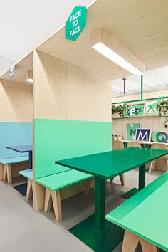 an empty table and benches in a room with green tables on either side, and blue chairs at the end