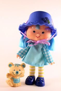 a doll with blue hair is standing next to a teddy bear and wearing a hat