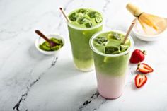 two cups filled with green smoothie and strawberries on a marble countertop next to other ingredients