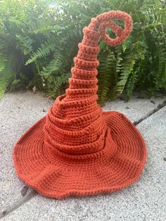 an orange crocheted hat sitting on the ground next to some bushes and plants