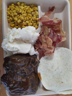 a white container filled with food on top of a wooden table next to tortillas and corn