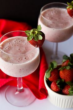 two glasses filled with milk and strawberries on top of a red cloth next to a bowl of strawberries