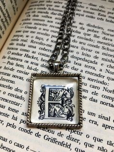 "Necklace with pendant made from a vintage book page, featuring an ornate letter \"E.\". This is a one-of-a-kind piece. It can be purchased with a silver-colored chain, with an organza ribbon with a metal clasp, or with both options for changing. No books were armed on purpose for the making of this necklace! The page used here was saved from a severely damaged book and given a new life as this unique piece." Vintage Silver Initial Pendant Necklace, Vintage Initials Pendant Necklace, Vintage Initials Necklace As Personalized Gift, Vintage Initials Necklace For Personalized Gift, Silver Vintage Necklace For Personalized Gift, E Initial Necklace, E Initial, Necklace With Pendant, Old Book Pages