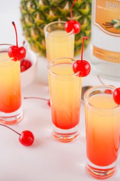 three glasses filled with liquid and cherries next to a pineapple drink in the background