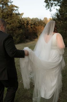 a bride and groom walking together in the grass with their veil pulled back over their shoulders