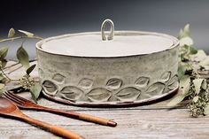 a wooden spoon sitting next to a white pot on top of a table with greenery