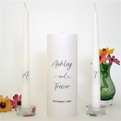 two white candles sitting next to each other on top of a table with flowers in vases