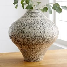 a vase sitting on top of a wooden table next to a window with green leaves in it