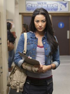 a woman standing in a hallway holding a folder and looking at the camera with an angry look on her face