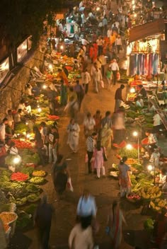 many people are walking around an outdoor market at night with lights on the ground and various fruits and vegetables being sold