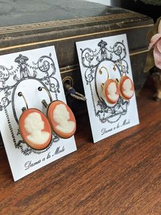 These are classical cameo styles!  A cream silhouette against a coral orange ground!  These are high-quality glass cameos. The cameos are available in two sizes: Larger 25mm or smaller 18mmThey are set in nickel-free leverback settings in your choice of Gold (smaller cameo shown in this finish), Antique Gold (larger cameo shown in this finish), Silver, or Antique Silver. Lots more Georgian- and Regency-inspired jewelry is available in my shop at Dames a la Mode: https://www.etsy.com/shop/damesal Cameo Earrings, Cameo Jewelry, Vintage Cameo, Leverback Earrings, Coral Orange, Inspired Jewelry, Earrings Vintage, Jane Austen, Vintage Earrings