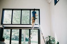 a man standing on a ladder painting the ceiling in a room with large windows and lots of plants