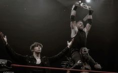 two men standing on top of each other in front of a wrestling ring holding their hands up