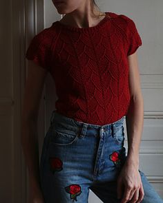 a woman wearing jeans and a red top with roses on the side, leaning against a wall