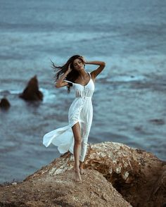 a woman standing on top of a rock next to the ocean wearing a white dress