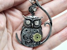 The Steampunk Owl Sitting on a Tree Branch Necklace is a captivating piece that marries the wisdom of nature with the intricate mechanics of steampunk design. The pendant features a detailed owl perched on a tree branch, its body adorned with gears and cogs that evoke a sense of retro-futuristic charm. The combination of organic and mechanical elements creates a striking visual contrast, making this necklace a standout accessory. Suspended on a 22-inch stainless steel chain, the pendant's cool m Fantasy Metal Necklaces For Collectors, Steampunk Style Chain Jewelry As Gift, Steampunk Metal Jewelry With Round Pendant, Steampunk Metal Necklace With Adjustable Chain, Steampunk Style Metal Necklace With Adjustable Chain, Steampunk Metal Round Pendant Jewelry, Steampunk Silver Round Pendant Jewelry, Steampunk Adjustable Necklace With Soldered Details, Steampunk Necklace With Adjustable Metal Chain