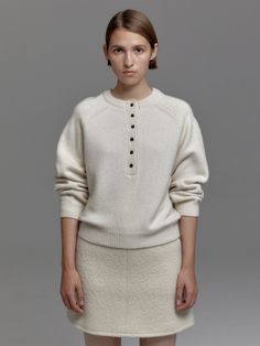 a woman standing in front of a gray background wearing a white sweater and beige skirt