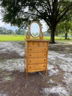 an old dresser with a mirror on it in the middle of a dirt field next to a tree