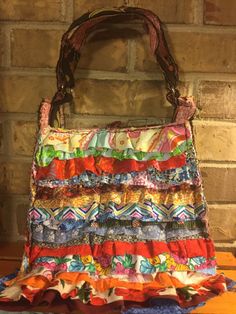 A handbag with multicolored ruffles on one side and a floral design on the other.  An unusual handle design where two handles are actually one. A button loop closure reveals a bright floral interior. Floral Interior, Top Handle Bags, Handle Design, Purses And Handbags, Top Handle, Ruffles, Reusable Tote Bags, Floral Design, Top Handle Bag