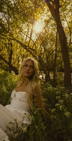 a woman in a white dress sitting on the ground next to some trees and bushes