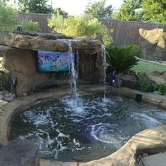 an outdoor hot tub with waterfall and rock walls, surrounded by landscaping items in the back yard