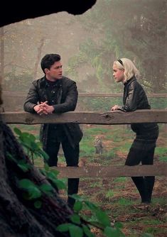 a man and woman standing next to each other near a wooden fence in the woods