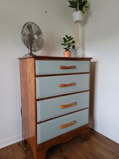 a blue dresser with three drawers and a fan next to it on top of a hard wood floor