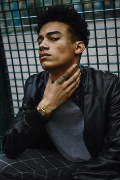 a man with his hands on his chest sitting in front of a metal fence and looking off to the side