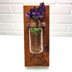 purple flowers are in a mason jar hanging on a wooden plaque