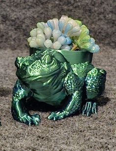 two green frog figurines sitting next to each other in front of a potted plant