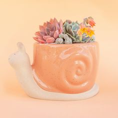 On a peachy background is an orange snail shaped planter filled with a cactus and succulent arrangement. Plants In Cute Pots, Cactus Planter Ideas Indoor, Mushroom Pinch Pot, Unique Clay Art, Fun Pottery Ideas, Kawaii Apartment, Pottery Glaze Ideas, Snail Planter, Novelty Planters