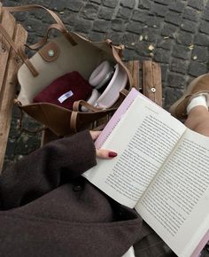 a woman reading a book while sitting on a bench with her purse and headphones