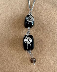 This pendant necklace has been made using 2 Obsidian gemstone, drilled cabochons, which have been decoratively wrapped with silver plated wire, embellished with a coiled, silver plated wire spiral and linked together to form a double length pendant. The base of the pendant has a smoky quartz pendant bead and the whole pendant is hung on a chunky, 3mm flat curb chain, with a lobster claw clasp. Pendant total length: 8cm Pendant width: 2.5cm Necklace length: 16"(40.5cm) Obsidian gemstones: approxi Elegant Onyx Wire Wrapped Necklace, Spiral Wire Wrapped Sterling Silver Necklace, Elegant Black Spiral Jewelry, Black Agate Wire Wrapped Jewelry, Spiral Black Necklace As A Gift, Silver Obsidian Necklaces, Black Spiral Necklace As A Gift, Spiral Wire Wrapped Necklace For Gift, Silver Obsidian Jewelry With Gemstone Beads