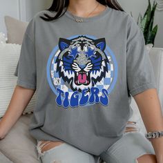 Get this popular new Tigers Mascot shirt and show off your school spirit in style!  Each shirt is printed in-house by me using high-quality materials on a Comfort Colors shirt, known for its exceptional comfort, style, and durability. Even the glitter is printed! No need to worry about peeling glitter vinyl, or messy glitter flakes. Available in unisex sizes S to 3X, and youth sizes, we've got you covered regardless of your preferred fit. The Preppy Tigers Shirt features a classic crew neck and short sleeves, making it suitable for year-round wear. The flattering silhouette ensures a comfortable and stylish fit for both men and women. Please note this listing is ONLY for comfort colors grey Note: As each shirt is individually printed, slight variations in color and placement may occur, add Casual College Shirt With Sublimation Print, Graphic Print Cotton Shirt For College, Blue Graphic Print Top, Blue School Spirit T-shirt With Sublimation Print, Blue School Spirit T-shirt With Heat Transfer Vinyl, Blue T-shirt With Sublimation Print For School Spirit, School Spirit Graphic Print Shirt For Streetwear, Team Spirit Graphic T-shirt For School, Team Spirit Graphic Print School T-shirt