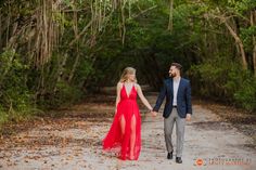 a man and woman holding hands walking down a dirt road in front of trees with leaves on the ground