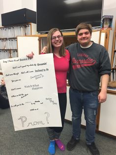 two people standing next to each other in front of a book shelf holding a sign