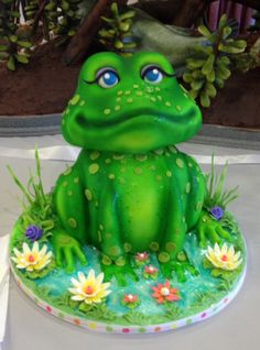 a green frog cake sitting on top of a white table with flowers and plants around it