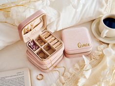 an open pink jewelry box sitting on top of a bed next to a cup of coffee