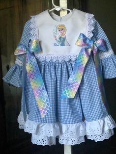 This dress is made from a pretty blue gingham   It is double skirted with a white underskirt and blue gingham over skirt. Both skirts are trimmed in white eyelet lace. The bib is made from a linen and has Elsa embroidered on it   The bib is trimmed with Venice lace and ties with blue and pink ribbons. This dress is available in sizes 1-8  the dress is priced at &45. The child's name can be embroidered on the dress or the bib.  The dress can be worn without the bib White Lace Trim Dress For Picnic, Cotton Lace Trim Dress For Picnic, Cotton Gingham Dress With Ruffle Hem, Gingham Cotton Dress With Ruffle Hem, Cute Gingham Dresses With Lace Trim, Spring Plaid Dress With Lace Trim, Cotton Gingham Dress With Lace Trim, Picnic Gingham Dress With Lace Trim, Gingham Cotton Dress With Lace Trim