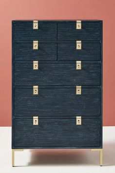 a blue chest of drawers with gold handles and knobs on each drawer, against a pink wall