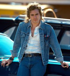 a man with long hair wearing a jean jacket and jeans is looking at the camera