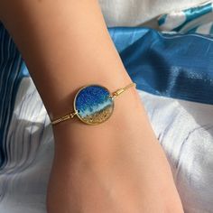 Dive into the depths of style with our ocean and beach-themed bracelet, a perfect accessory for those who adore the sea.  Adjustable up to 20 cm in length, this bracelet features a 22 mm diameter ring adorned with a captivating oceanic and beach view. Crafted with meticulous attention to detail, all metal parts are 24K gold-plated, ensuring both durability and elegance. Handcrafted with resin and resin pigments, each piece is unique, embodying the serene beauty of the ocean. Whether you're a beachcomber or a sailor at heart, this bracelet is sure to make a splash. Packaged with care, it makes for an ideal gift for any ocean lover. Let the waves of style wash over you or surprise a loved one with a piece of the sea. Order now and let the adventure begin! Due to the nature of handmade produc Ocean-inspired Turquoise Bracelets As Gift, Turquoise Ocean-inspired Bracelets For Gift, Turquoise Ocean-inspired Bracelets As Gift, Beachy Sand-colored Jewelry For Gift, Beachy Sand-colored Jewelry Gift, Blue Bracelets With Lobster Clasp For Beach, Ocean-inspired Bracelet Jewelry For Beach Season, Ocean-inspired Strand Friendship Bracelets As Gifts, Ocean-inspired Strand Friendship Bracelets As Gift