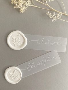 two clear plastic labels with white writing on them next to some dried flowers and branches