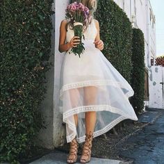 a woman in a white dress is holding flowers and standing next to a wall with ivy growing on it