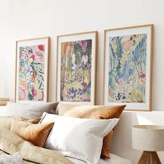 three framed paintings hang on the wall above a bed with white sheets and beige pillows